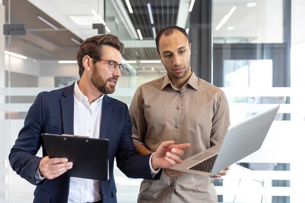 Two male colleagues review information on a laptop, collaborating inside an office setting for a business discussion.
